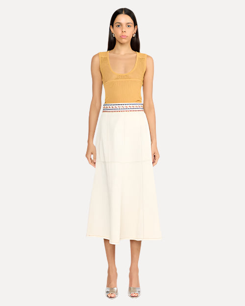 A dark-haired model wearing a sleeveless mustard yellow top with a scoop neckline, tucked into a white midi skirt with an embroidered blue and red waistline. The model is wearing silver open-toed heels to match and is standing in front of a white background.