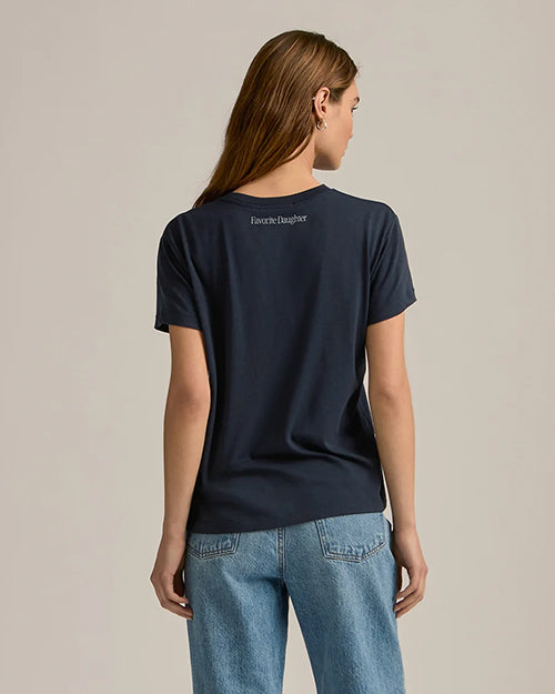 A backside view of a brunette model with her hair swept to the side, wearing a short-sleeve navy crewneck t-shirt. Under the neck trim reads "Favorite Daughter". The modelis also wearing light blue jeans.