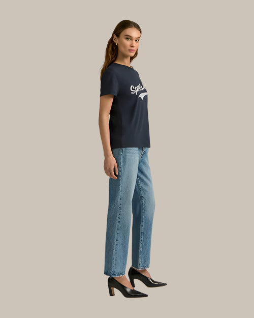 A side view of a person wearing a navy-blue t-shirt with the text "Sports Mom" written in white across the chest in vintage baseball font. The text is in a bold, uppercase font and is centered on the chest area. The t-shirt has a classic crew neckline and short sleeves. The fabric appears to be soft and slightly stretchy, providing a comfortable fit. The person is also wearing light blue jeans and black high heels.
