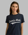 An upper body shot of a stylish brunette model wearing a short-sleeve, navy crewneck t-shirt with the text "Sports Mom" written in white across the chest in vintage baseball font, with gold hoop earrings. She is standing against a grey background.