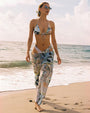 A model wearing a bikini with a floral/paisley print and a slightly transparent matching sarong and drawstring pants. The composition places the woman against a background of ocean water and sky, with a clear horizon line. Colors are predominantly pastel blues, tans, and whites.