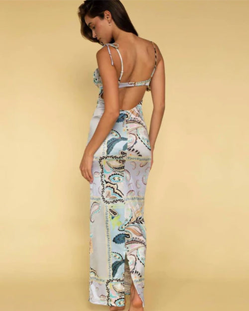 A full body backside shot of a barefoot brunette model standing against a yellow background, wearing a long form-fitting dress with thin straps and shoulder bows, featuring an open back with a strip in the middle, and a colorful pattern with various paisley designs.