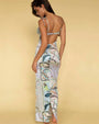 A full body backside shot of a barefoot brunette model standing against a yellow background, wearing a long form-fitting dress with thin straps and shoulder bows, featuring an open back with a strip in the middle, and a colorful pattern with various paisley designs.