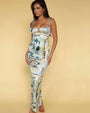 A full body shot of a barefoot brunette model standing against a yellow background, wearing a long form-fitting dress with thin straps and shoulder bows, featuring an empty space design at the chest, and a colorful pattern with various paisley designs.