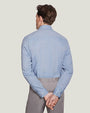 A knee-up backside view of a model wearing a light-blue button up shirt tucked into gray buttoned pants, standing in front of a white background.