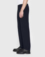 A lower body sideview of a model wearing a white shirt tucked into navy trousers, and black boots. The background is plain and light-colored.