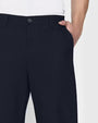 A close-up frontal view of a models' hip with his hand in his pocket, wearing a white shirt tucked into navy trousers. The background is plain and light-colored.
