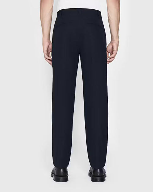 A lower body backside view of a model wearing a white shirt tucked into navy trousers, and black dress shoes. The background is plain and light-colored.