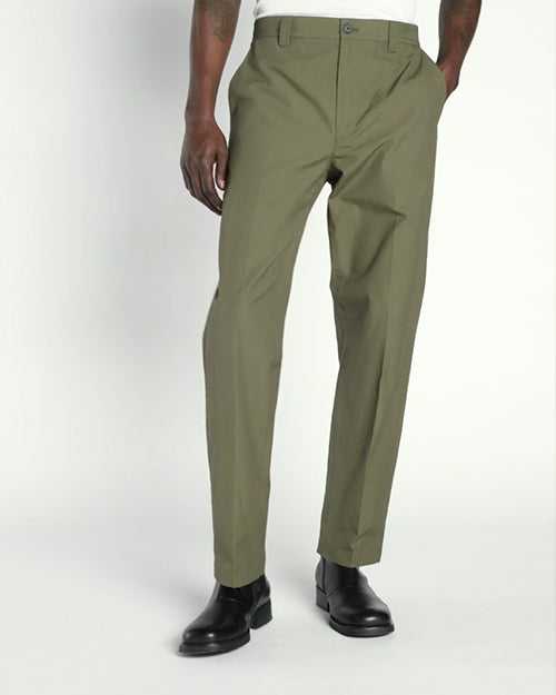 A lower body view of a model with his hand in his pocket, wearing a white shirt tucked into green trousers and black dress shoes. The background is plain and light-colored.