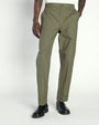 A lower body view of a model with his hand in his pocket, wearing a white shirt tucked into green trousers and black dress shoes. The background is plain and light-colored.