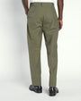 A lower body backside view of a model with his hand in his pocket, wearing a white shirt tucked into green trousers, and black dress shoes. The background is plain and light-colored.