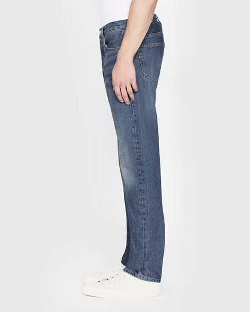 A lower body sideview of a model wearing blue jeans with a faded look tucked into a white t-shirt and sneakers. The background is completely white.