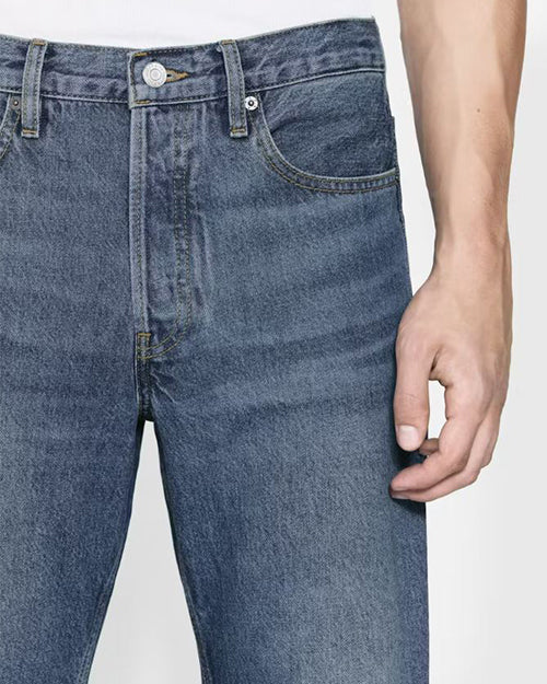 A close-up waist level view of a model wearing blue jeans with a slightly faded look, tucked into a white shirt. The background is plain white.