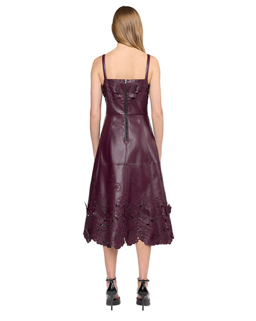 Back view of a person wearing a sleeveless dark purple leather dress with thin straps, a center back zipper, and floral cut-out detailing along the hem, paired with black ankle-strap high heels.