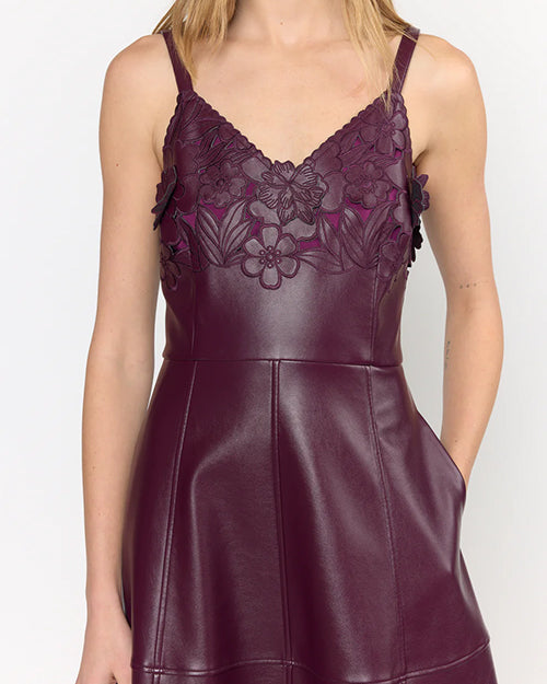 Close up of a person wearing a sleeveless dark purple dress with floral embroidery on the bodice, thin straps, V-neckline, and a smooth leather-like skirt, with one hand in a side pocket