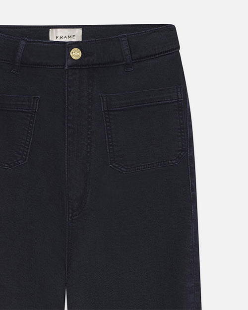 A closeup front view of a pair of navy denim jeans with a gold button. The jeans have zipper and button closure, two large front pockets, and a label reading FRAME.