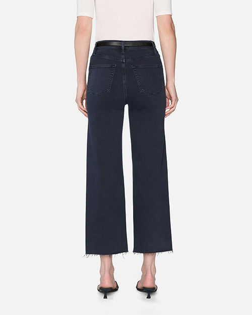 The backside view of a model wearing a white short-sleeve top tucked into ankle-length navy denim jeans with a black belt and black open-toed heels. The jeans have back pockets and frayed hems at the bottom.