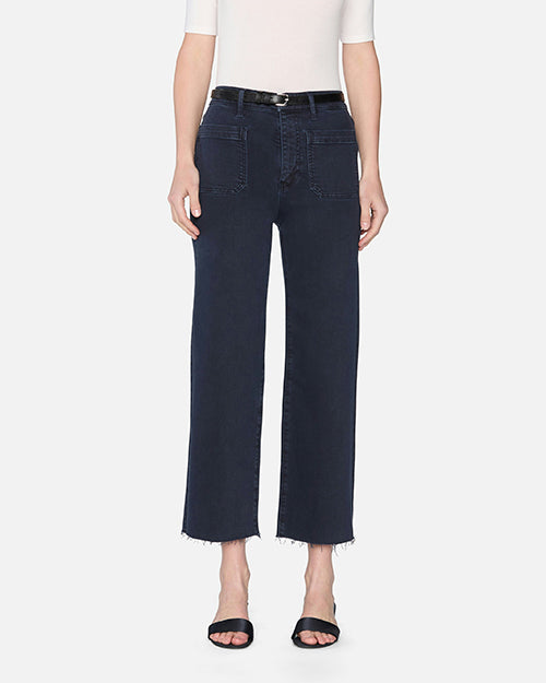 A model wearing a white short-sleeve top tucked into ankle length navy denim jeans with a black belt and black open-toed heels. The jeans have zipper and button closure, two large front pockets, and frayed hems at the bottom.