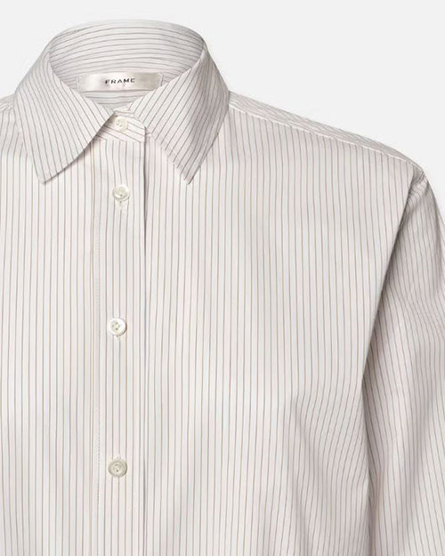 A close-up shoulder level image of a white long-sleeve button-up shirt with a collar. The shirt has thin vertical stripes and white buttons. The inside neckline contains a tag that reads "FRAME".