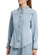 A torso shot of a brunette model wearing a light blue collared denim shirt with long sleeves and long wrist cuffs. The shirt features visible stitching along the seams and silver buttons, and the background is plain white.