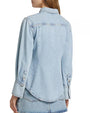 The backside view of a brunette model with her hair swept to the side, wearing a collared light blue denim shirt with long sleeves, long wrist cuffs and visible stitching along the seams and buttons.