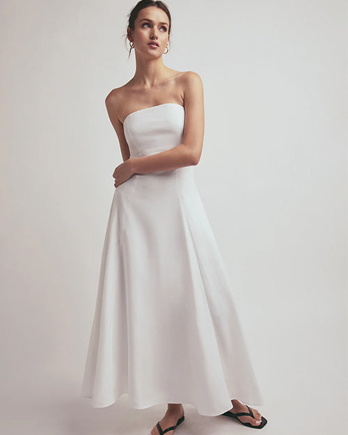 A full body shot of a brunette model with her hand wrapped around her waist and her hair tied up wearing a long, strapless white dress that reaches down to the ankles. The model is also wearing a pair of gold hoop earrings, black flip-flops and is standing in front of a white background.
