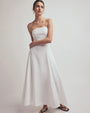 A full body shot of a brunette model with her hand wrapped around her waist and her hair tied up wearing a long, strapless white dress that reaches down to the ankles. The model is also wearing a pair of gold hoop earrings, black flip-flops and is standing in front of a white background.