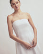 A waist-up shot of a brunette model with her hair up wearing a long, strapless white dress, and a pair of gold hoop earrings in front of a white background.