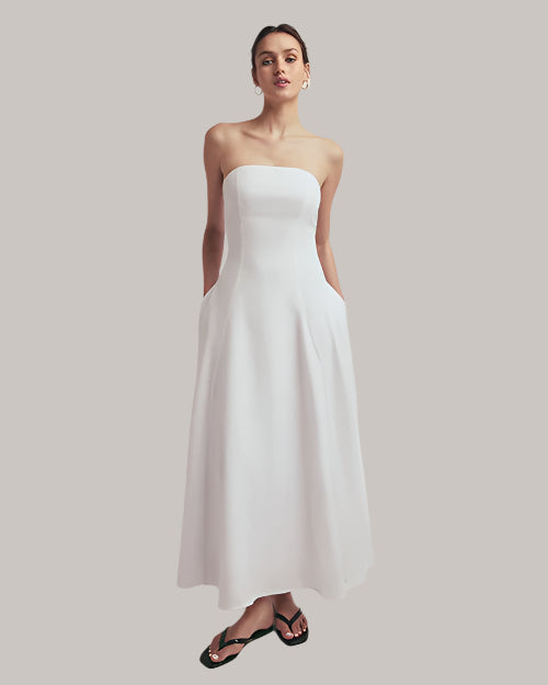 A full body shot of a brunette model with her hair up wearing a long, strapless white dress that reaches down to the ankles. The model is also wearing a pair of gold hoop earrings, black flip-flops and is standing in front of a white background.