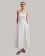 A full body shot of a brunette model with her hair up wearing a long, strapless white dress that reaches down to the ankles. The model is also wearing a pair of gold hoop earrings, black flip-flops and is standing in front of a white background.