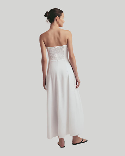 The backside view of a brunette model with her hair tied up wearing a long, strapless white dress with a mostly open back with an elastic material and a zipper. The model is also wearing black flip-flops and is standing against a plain background.