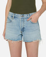 A waist-level image of a model with her hand in her pocket, wearing a green top tucked into light-wash denim shorts with frayed ends. The model is standing in front of a white background.