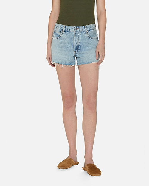 A frontal lower body image of a model wearing a green top tucked into light-wash denim shorts with frayed ends, and brown sandals. The model is standing in front of a white background.