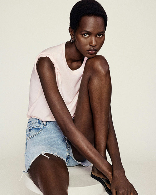 A model sitting on a white chair in a white room with one leg bent and one extended. The person is wearing sleeveless light pink top, light-wash denim shorts with frayed ends, and black flip flops.