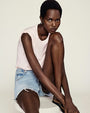 A model sitting on a white chair in a white room with one leg bent and one extended. The person is wearing sleeveless light pink top, light-wash denim shorts with frayed ends, and black flip flops.