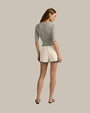 A backside view of a brunette model with her hair swept to the side, wearing a grey, short-sleeved top with a round neckline tucked into white athletic track style shorts with green trim along the sides and hem, an elastic waistband with two green stripes. The model is also wearing black sandals with a simple strap design. The background is plain and grey.