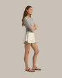 A sideview of a brunette model wearing a grey, short-sleeved top with a round neckline tucked into white athletic track style shorts with green trim along the sides and hem, an elastic waistband with two green stripes. The model is also wearing black sandals with a simple strap design. The background is plain and grey.