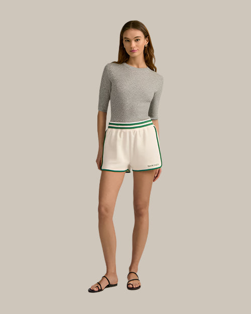 A brunette model wearing a grey, short-sleeved top with a round neckline tucked into white athletic track style shorts with green trim along the sides and hem, an elastic waistband with two green stripes, which read "Favorite Daughter" at the bottom of the left leg in green text. The model is also wearing black sandals with a simple strap design. The background is plain and grey.