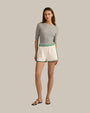A brunette model wearing a grey, short-sleeved top with a round neckline tucked into white athletic track style shorts with green trim along the sides and hem, an elastic waistband with two green stripes, which read "Favorite Daughter" at the bottom of the left leg in green text. The model is also wearing black sandals with a simple strap design. The background is plain and grey.
