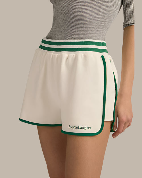 A waist-level view of a model wearing a grey, short-sleeved top tucked into white athletic track style shorts with green trim along the sides and hem, an elastic waistband with two green stripes. The shorts read "Favorite Daughter" at the bottom of the left leg in green text. The background is grey.