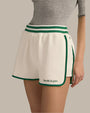 A waist-level view of a model wearing a grey, short-sleeved top tucked into white athletic track style shorts with green trim along the sides and hem, an elastic waistband with two green stripes. The shorts read "Favorite Daughter" at the bottom of the left leg in green text. The background is grey.