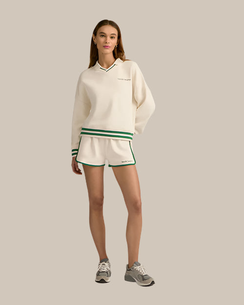 A brunette model standing against a plain background, wearing a white long-sleeve sweater with small font on the left chest, green trim on the collar, waistband and cuffs, paired with matching white athletic style shorts with green trim along the sides and hem. The shorts have an elastic waistband with 2 green stripes, and the person is also wearing gray athletic shoes.