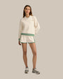 A brunette model standing against a plain background, wearing a white long-sleeve sweater with small font on the left chest, green trim on the collar, waistband and cuffs, paired with matching white athletic style shorts with green trim along the sides and hem. The shorts have an elastic waistband with 2 green stripes, and the person is also wearing gray athletic shoes.