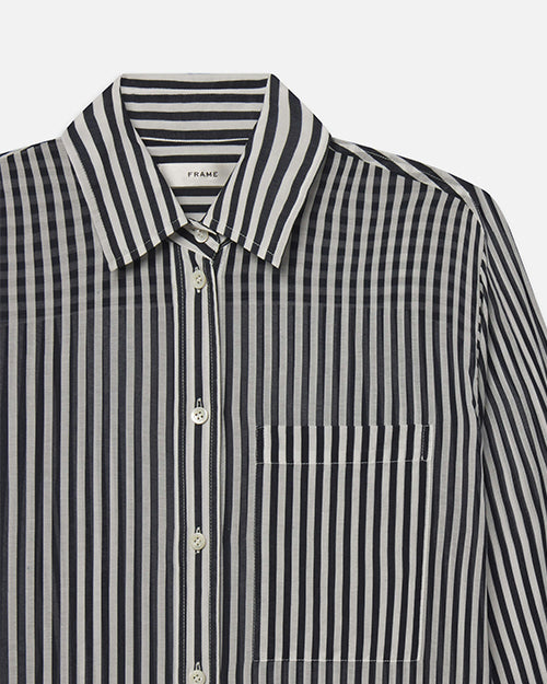 A close-up chest level view of a long-sleeved, button-up shirt with thin navy and white stripes, and a front pocket. The inner neckline has a white tag reading "FRAME".