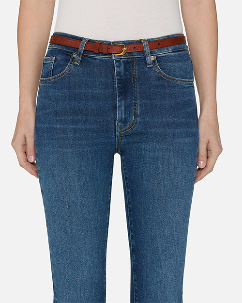 A close-up waist-level view of a model wearing blue jeans, a tucked in white top, and a brown belt and sandals. She is standing in front of a white background.