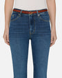 A close-up waist-level view of a model wearing blue jeans, a tucked in white top, and a brown belt and sandals. She is standing in front of a white background.