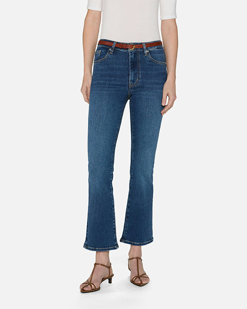 A lower body view of a model wearing blue jeans with a slight flare at the end, a tucked in white top, and a brown belt and sandals. She is standing in front of a white background.