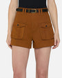 A waist-level view of a model wearing a black top tucked into high-waisted brown shorts with two large front pockets featuring flap closure and black buttons, and a thin black belt held by double belt loops.