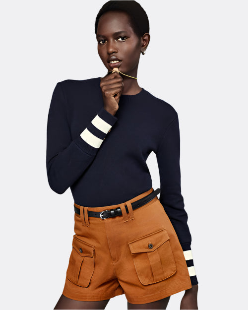 A model holding a gold necklace up to her chin, while wearing a black long-sleeve top with white stripes on the cuffs, tucked into high-waisted brown shorts with two large front pockets featuring flap closure and a thin black belt held by double belt loops.