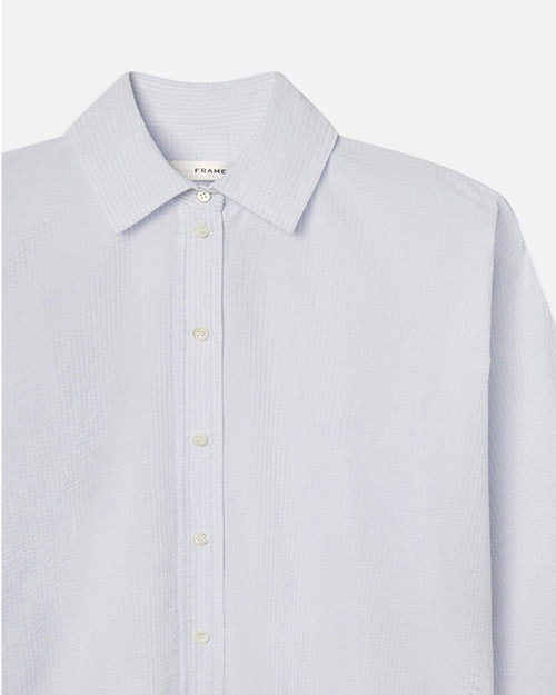 A close-up chest level view of a white and light blue striped long-sleeve button-up shirt. The inside neckline contains a tag that reads "FRAME".
